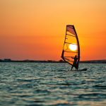 Seascape of still sea surface, man practicing wind surfing and golden sunset in sky on summer clear day. Still landscapes of travels and destination scenics