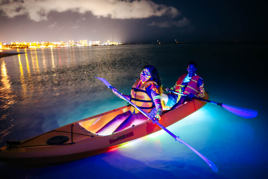 Two guests paddling an LED-lit clear kayak at night with the Aruba shoreline glowing behind them